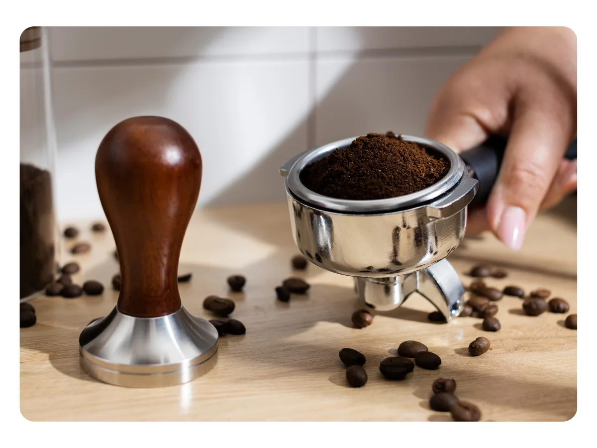 Close-up de um bastão de compactação de pó e o caximbo de uma máquina expresso com o pó, além de grão espalhados na bancada.